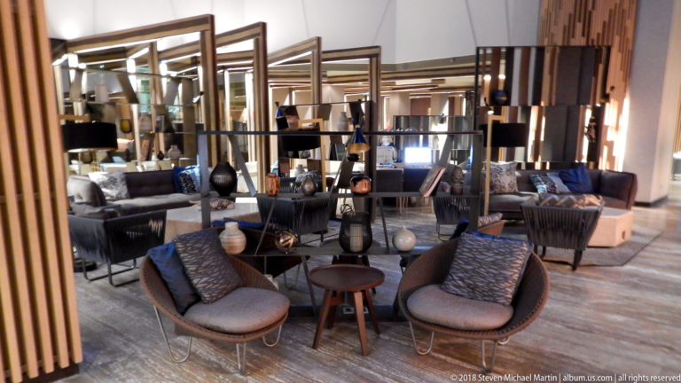 Hotel lounge with curved chairs, patterned cushions, open shelving, lamps, ceramics, and seating beyond.