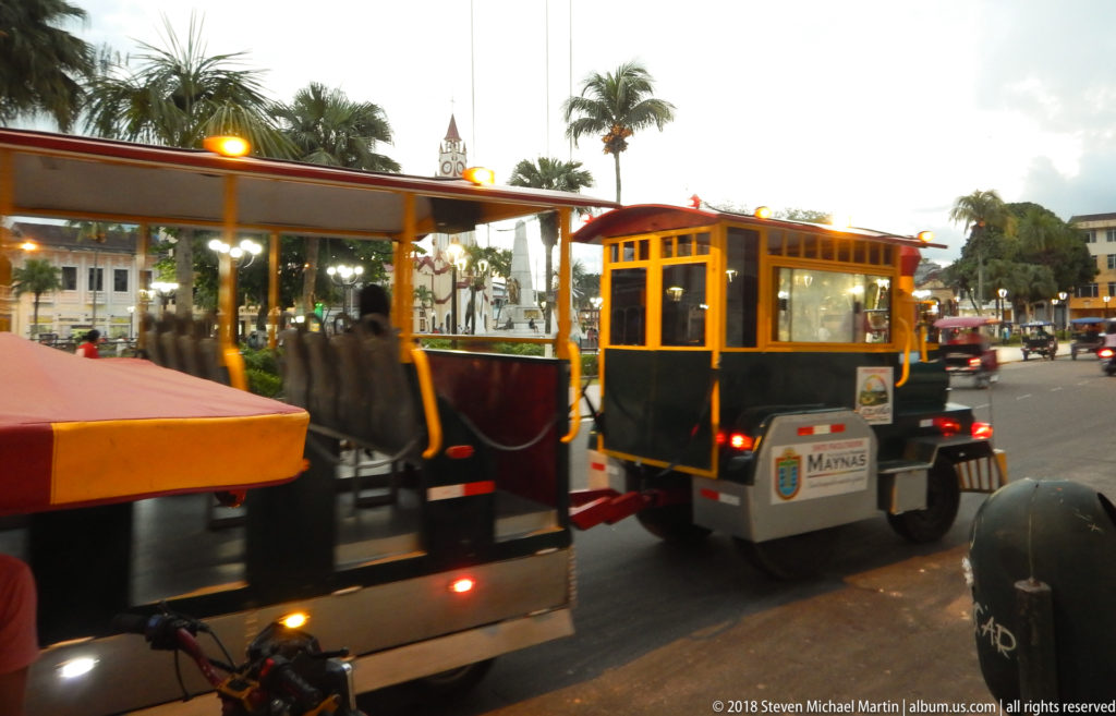 Orange-and-green municipal tourist road train marked Maynas traveling around a palm-lined city square.