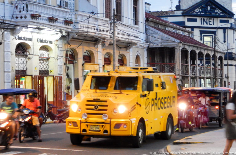 Yellow Prosegur armored truck moving through evening traffic with mototaxis and motorcycles.