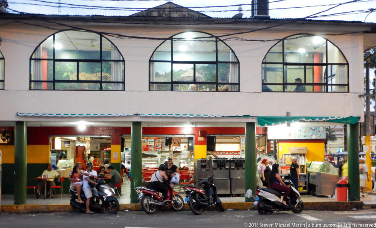 Open-front restaurant with diners below three large arched windows and motorcycles parked along the curb.