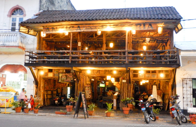 Rustic two-story café or restaurant with an open timber balcony, warm lights, potted plants, and parked motorcycles.