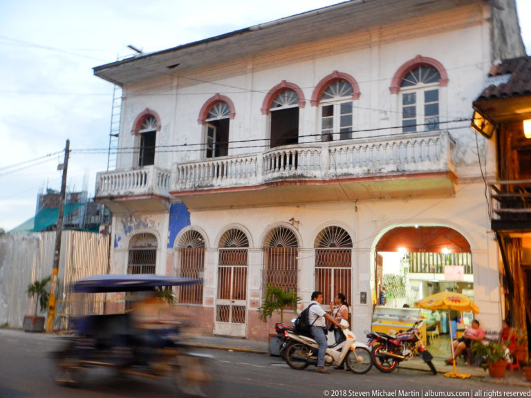 White two-story historic façade with red-trimmed arched windows, long balustraded balcony, and passing mototaxi.