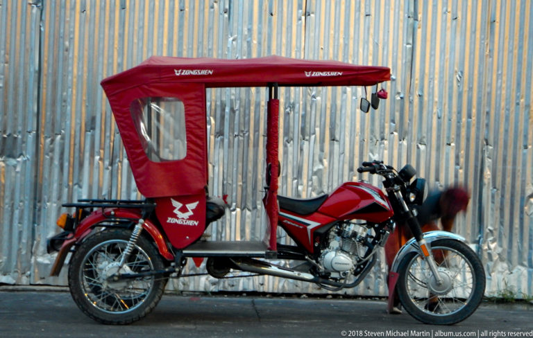 Red mototaxi in side profile parked against a corrugated-metal wall.