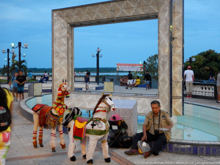 Two decorated ponies and a seated handler beside a riverside plaza and open waterfront.