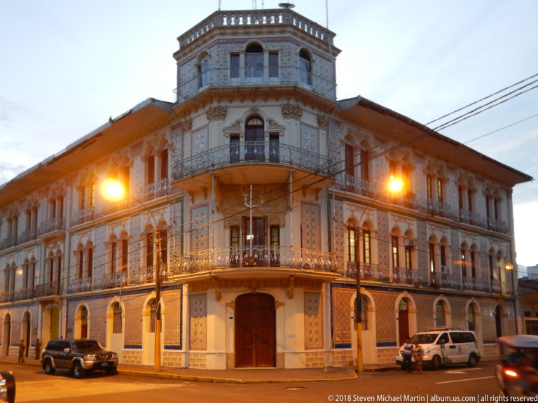 Ornate multi-story corner building with iron balconies, arched openings, patterned façade, and square rooftop pavilion.