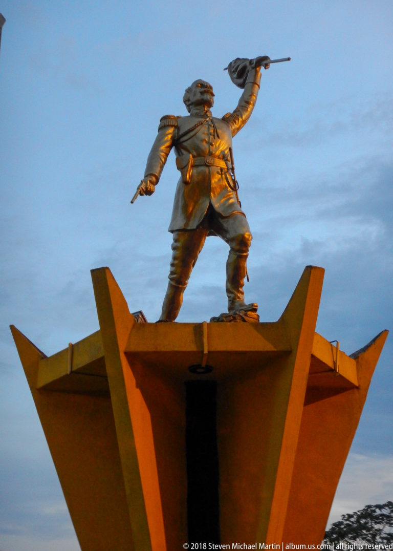 Bronze-colored standing figure pointing outward from a tall angular orange pedestal at dusk.