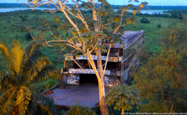 Elevated wooden lookout building among trees, with exterior stairs and a distant river or wetland beyond.