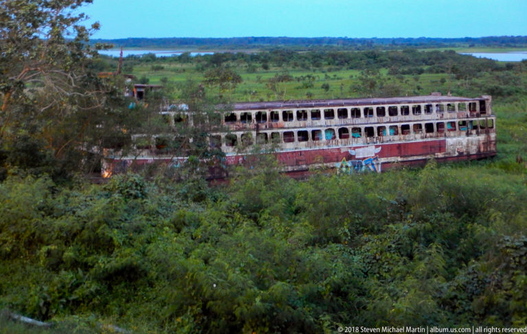 Large weathered multi-deck passenger vessel surrounded by dense green vegetation near a distant river.