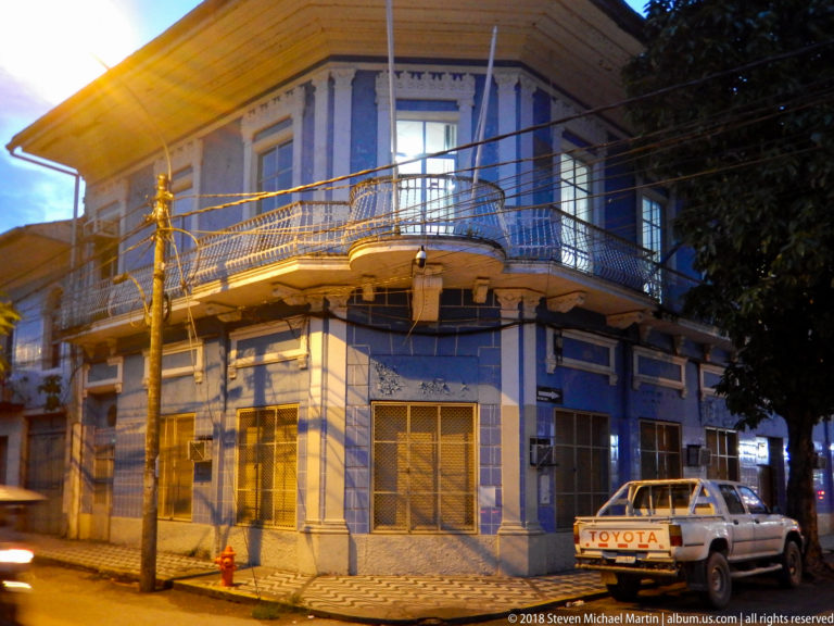 Blue-and-white two-story corner building with wraparound iron balcony, decorative trim, utility wires, and a pickup truck.