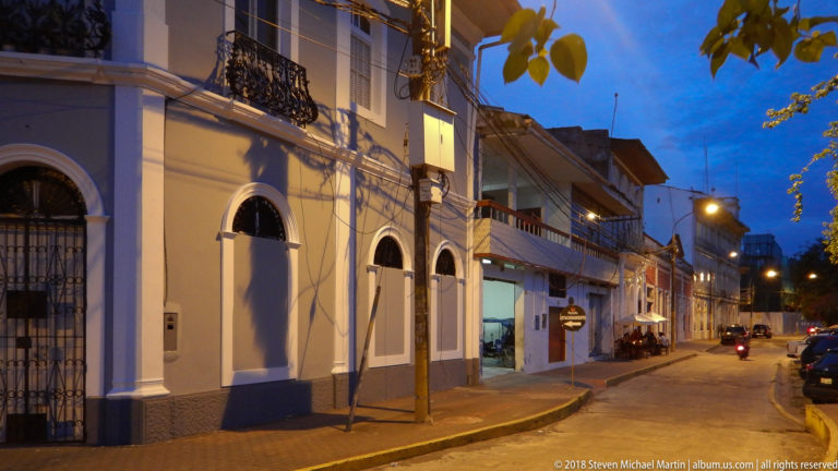 Pale arched façade and adjoining buildings along a quiet Iquitos street at dusk under warm street lamps.