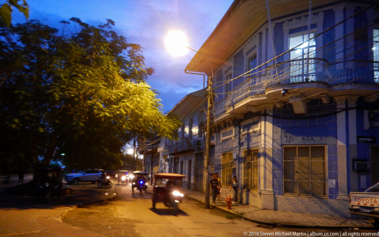 Mototaxi passing a blue-and-white historic corner building on a tree-lined Iquitos street at dusk.