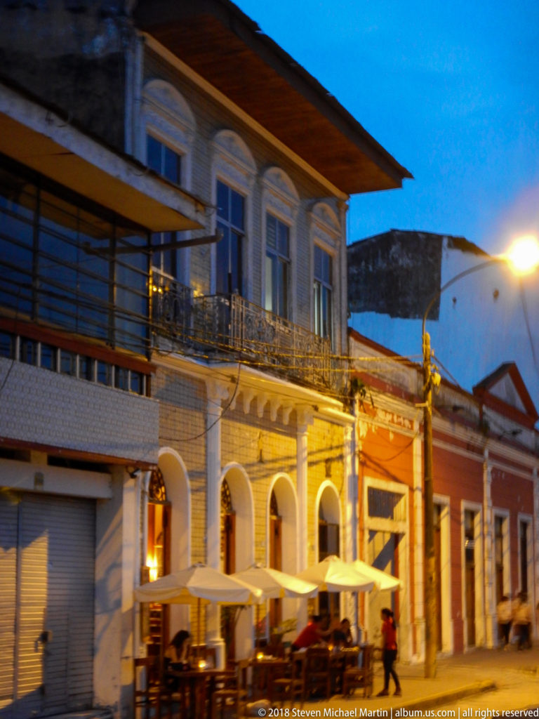 Two-story historic building with arched ground floor, iron balcony, broad roof, and sidewalk tables under white umbrellas.