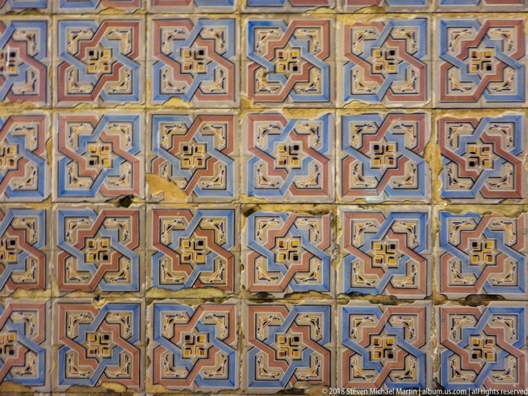 Close view of repeating blue, ochre, cream, and red geometric ceramic tiles on a historic façade.