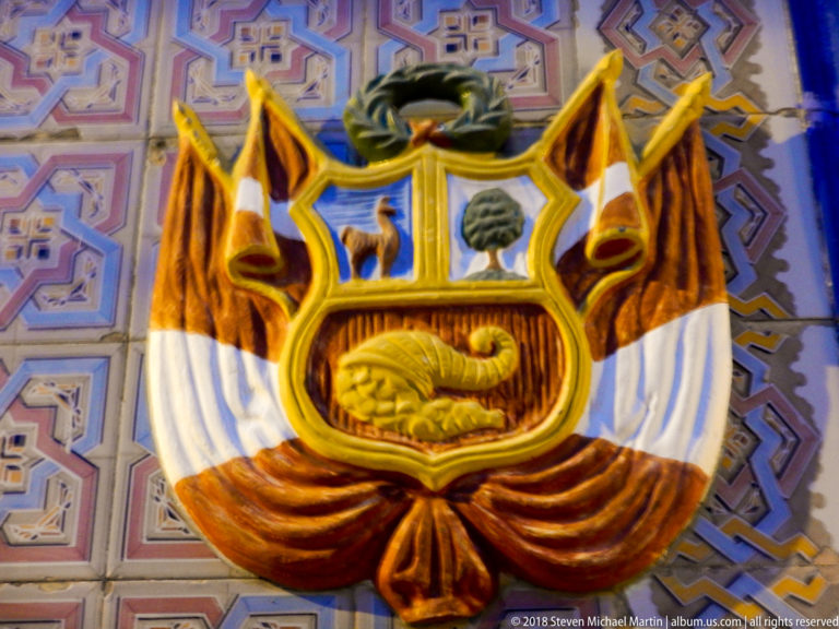 Relief of Peru’s coat of arms with vicuña, cinchona tree, cornucopia, flags, and wreath on patterned tiles.