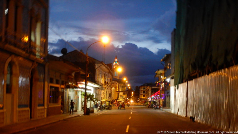 Long Iquitos street at blue hour with glowing lamps, low historic façades, pedestrians, and motorcycles.