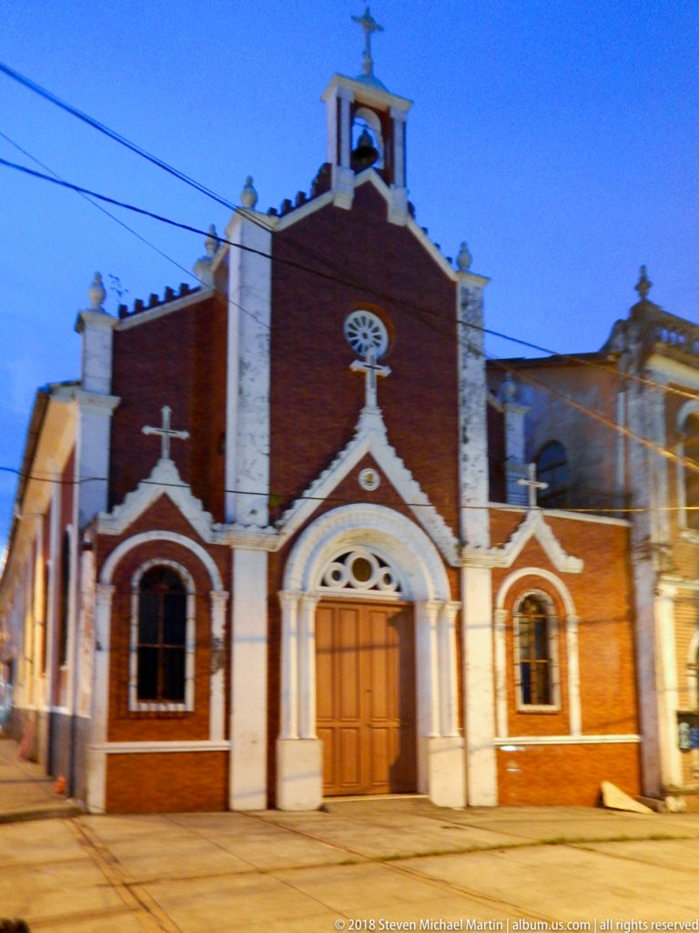 Small red-brick church with white-trimmed arched door and windows, circular gable details, and bell opening.