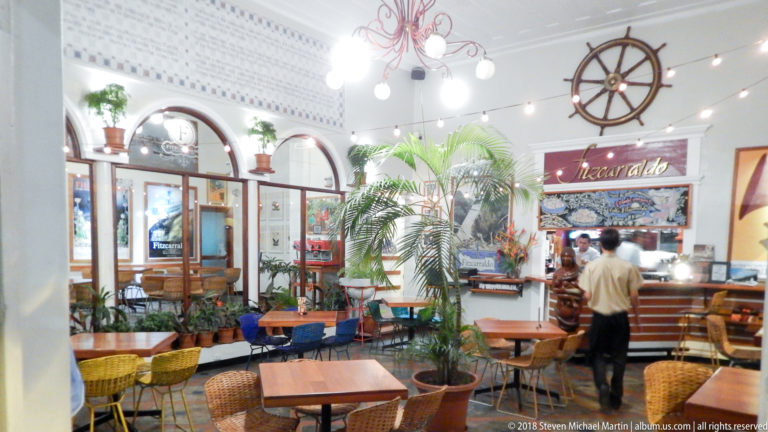 Bright restaurant interior with woven chairs, wooden tables, potted palms, arched openings, and a large ship’s wheel.