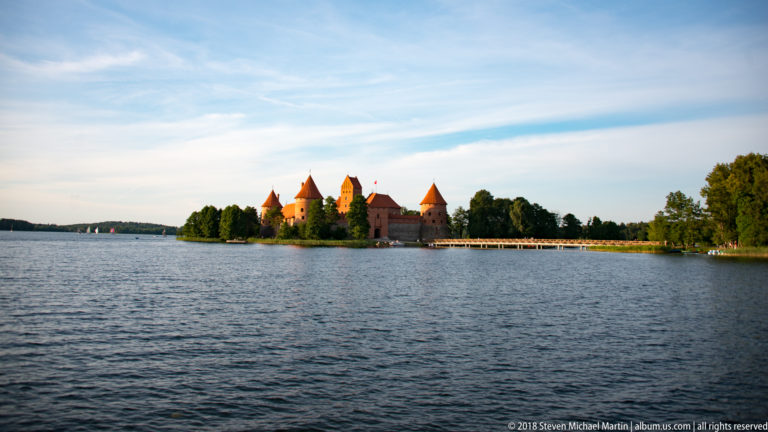 Distant Trakai Island Castle across a broad expanse of Lake Galvė, with trees and a long footbridge extending from shore.