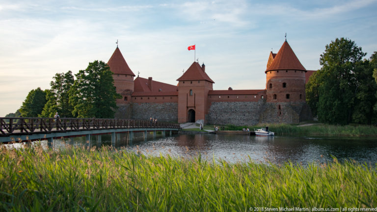 Trakai Island Castle across Lake Galvė with its wooden footbridge, red-brick towers, and reeds in the foreground.