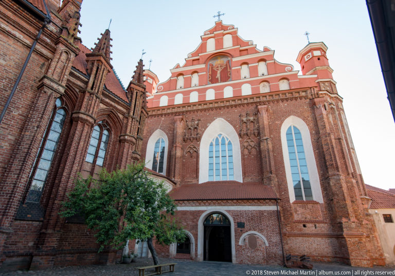 Red-brick Bernardine Church elevation with tall pointed windows, stepped gable, corner turrets, and open doorway.