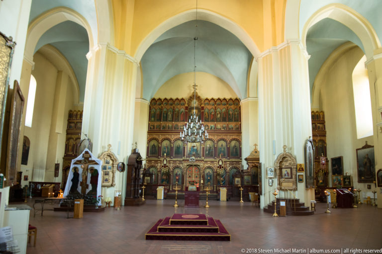 Wide Orthodox cathedral nave with tall white piers, red platform, side shrines, and iconostasis at far end.