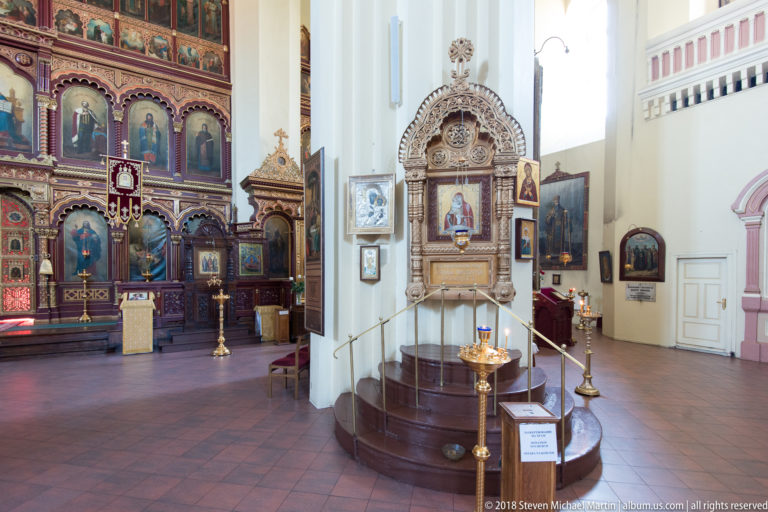 Raised wooden side shrine with icon, steps, brass lamp stand, and main iconostasis visible to the left.