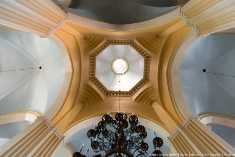 Upward view of cream ribs and pale vaults converging on an octagonal glazed lantern above a dark chandelier.
