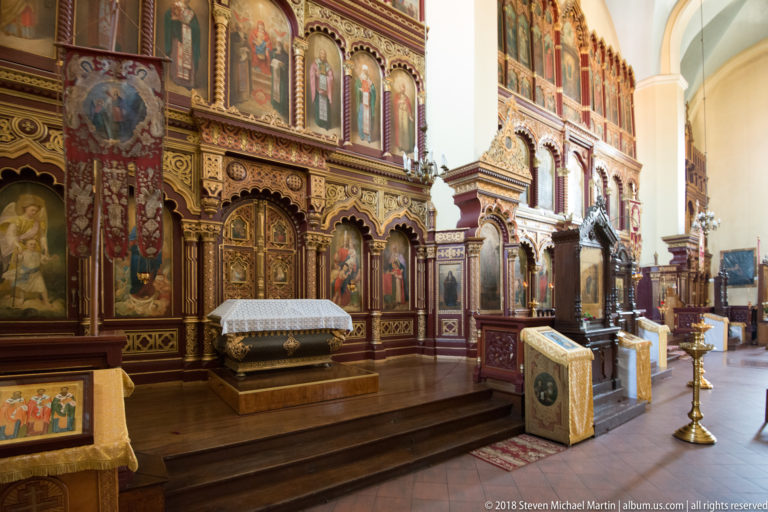 Oblique view of carved wooden iconostasis with painted saints, gilded details, lecterns, and brass candle stand.