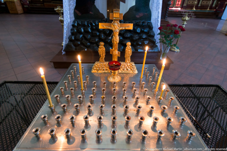 Metal votive stand with burning taper candles, a gilded crucifix group, flowers, and dark monument behind.