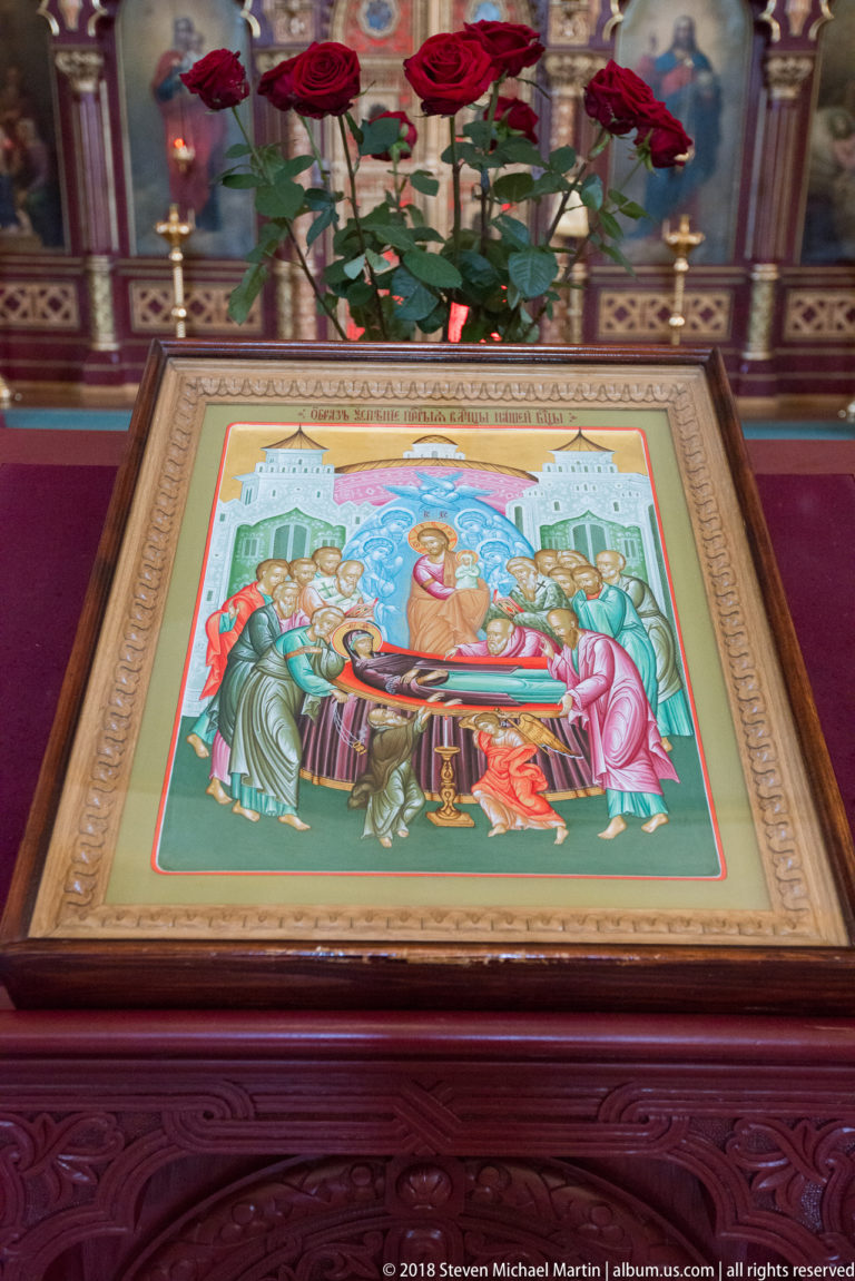 Orthodox icon on a sloped wooden stand, with red roses, candle stands, and ornate iconostasis behind it.