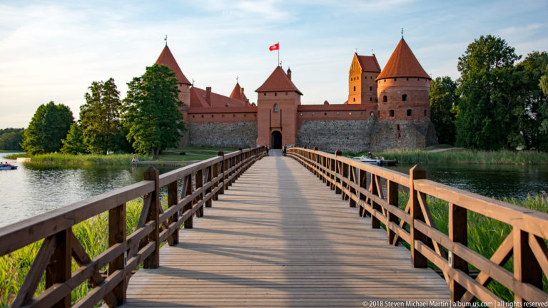 Wooden footbridge crossing Lake Galvė toward the red-brick gate and towers of Trakai Island Castle.