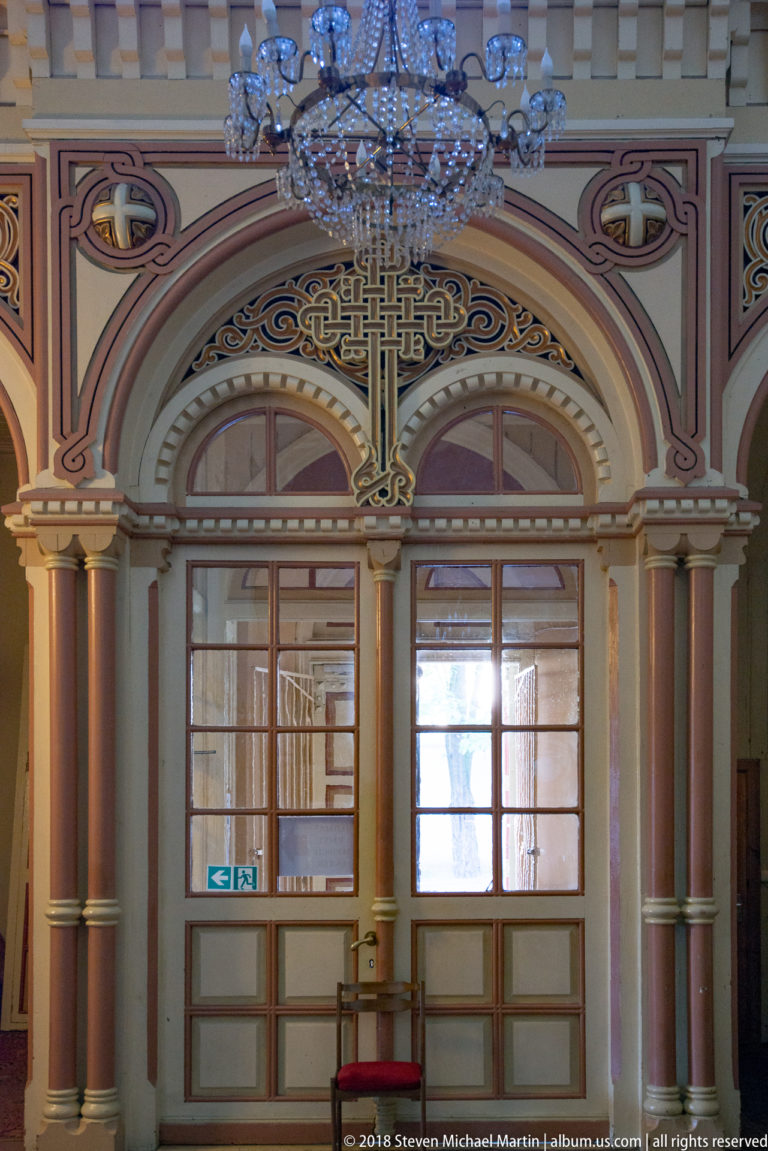 Decorated cathedral entrance bay with arched openings, slender columns, chandelier, patterned walls, and glazed double doors.