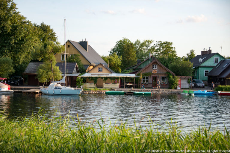 Wooden lakeside houses with gardens, sailboats, and small boats moored along calm water in Trakai.