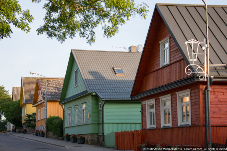 Row of colorful wooden houses with steep roofs, timber siding, and small gardens along a quiet Trakai street.