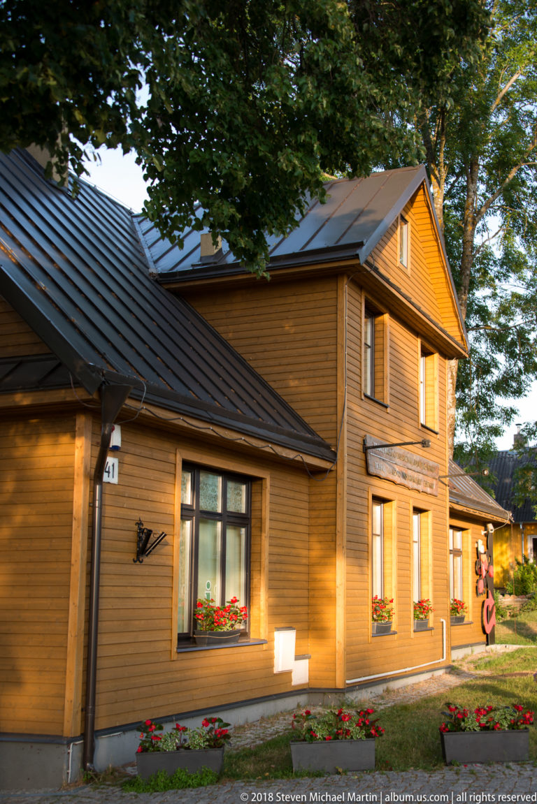 Two-story wooden house with brown siding, metal roof, flower boxes, and white-trimmed windows in warm evening light.