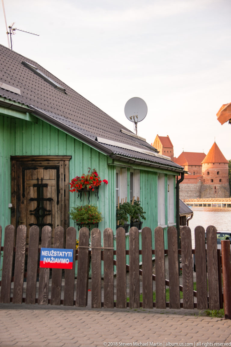 Green wooden house with shuttered windows, flowers, and picket fence in Trakai, with castle towers visible behind.