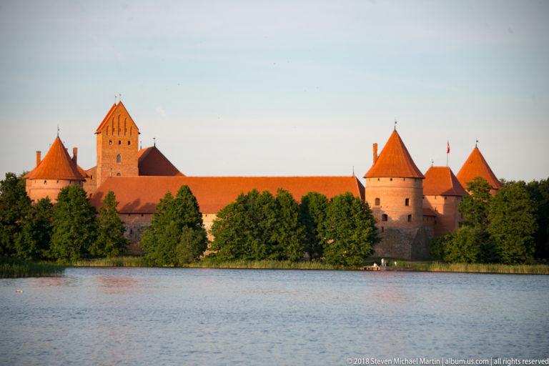Red-brick Trakai Island Castle with cylindrical towers and steep tiled roofs across Lake Galvė in evening light.