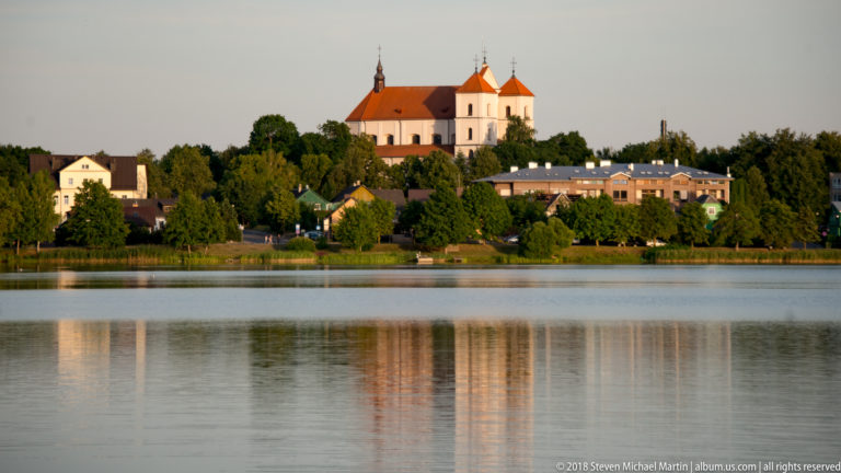 Pale church with red roof rising above a tree-lined Trakai lakeshore, reflected in calm water at evening.