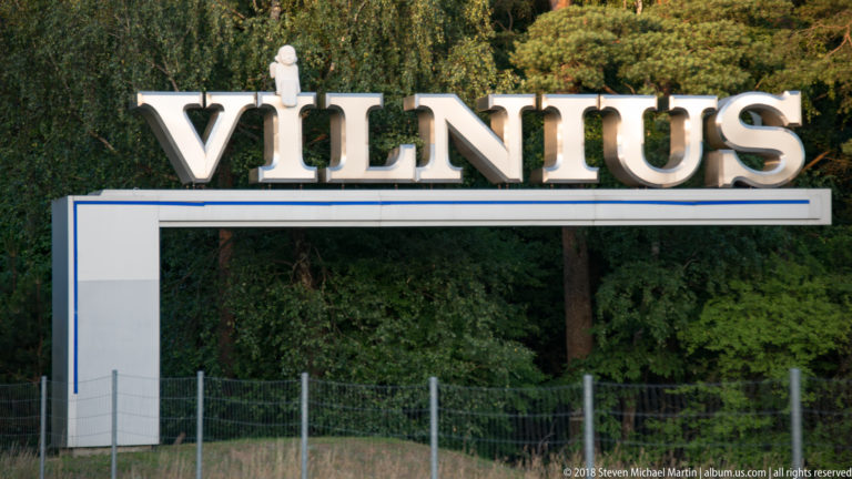 Large white letters spelling VILNIUS mounted on a rectangular frame with a small figure above, beside trees.