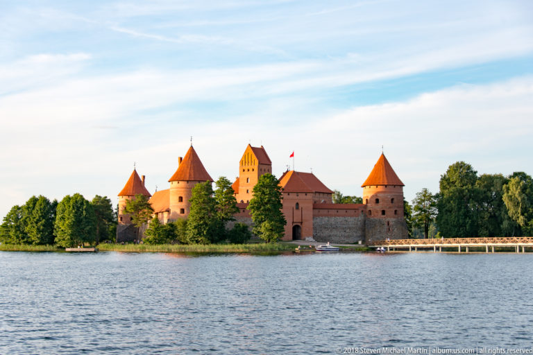 Trakai Island Castle across Lake Galvė in warm evening light, with red-brick towers, walls, and trees.
