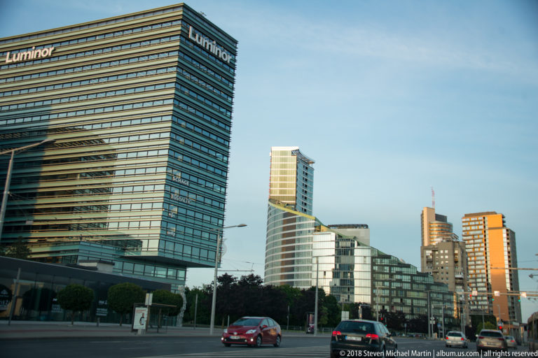Vilnius business district with glass high-rises, varied modern towers, roadway traffic, and a broad avenue under clear sky.