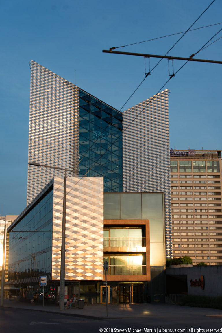 Modern glass-and-metal commercial complex with patterned high-rise façades, angular low building, and trolley wires overhead.