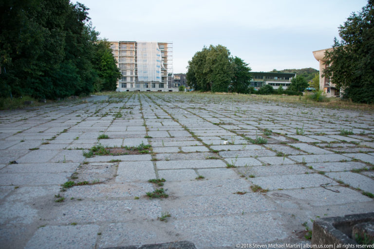 Wide cracked concrete plaza with weeds between slabs, trees at the edges, and a modern high-rise in the distance.
