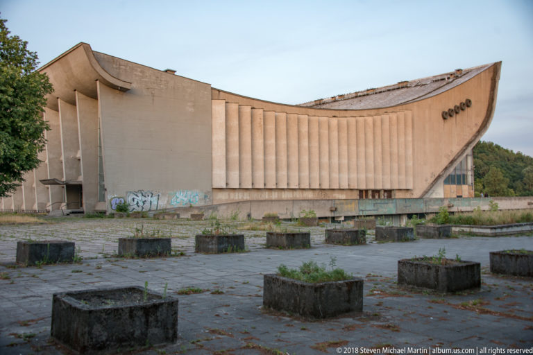 Large weathered concrete modernist building with curving roofline, vertical fins, graffiti, weeds, and empty planters.