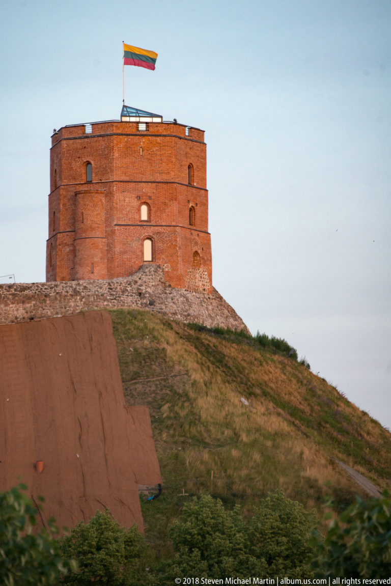 Red-brick Gediminas’ Tower on Castle Hill with Lithuanian flag flying above the roof and stone defenses below.