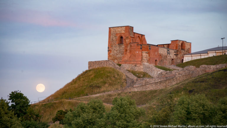 Ruined brick and stone walls of the Upper Castle on grassy Castle Hill with a bright moon rising beside the slope.