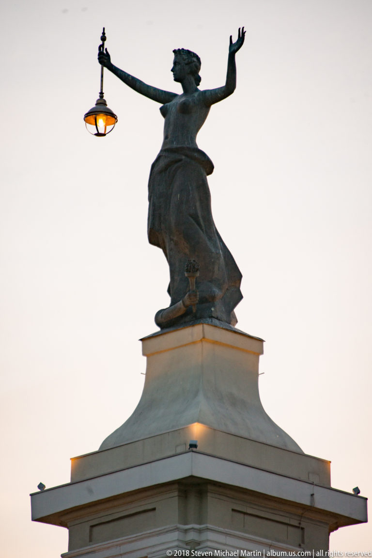 Bronze female rooftop figure in flowing drapery with arms extended above a pale classical building at dusk.