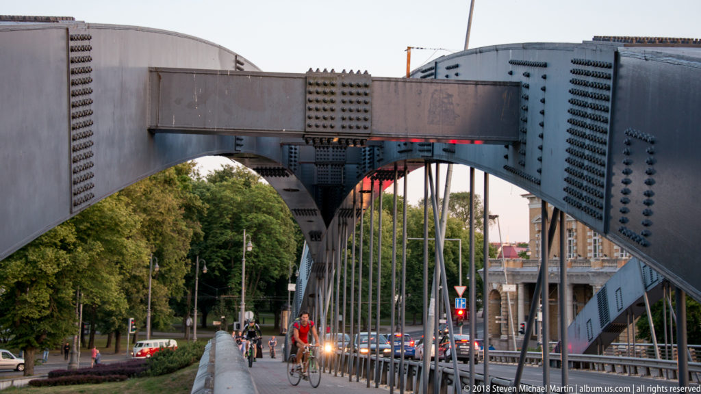 Riveted steel arch bridge with vertical suspenders, cyclists, pedestrians, and traffic crossing in central Vilnius.