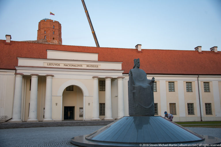 Lithuanian National Museum façade with classical columns, arched entrance, red roof, and a modern monument in the foreground.