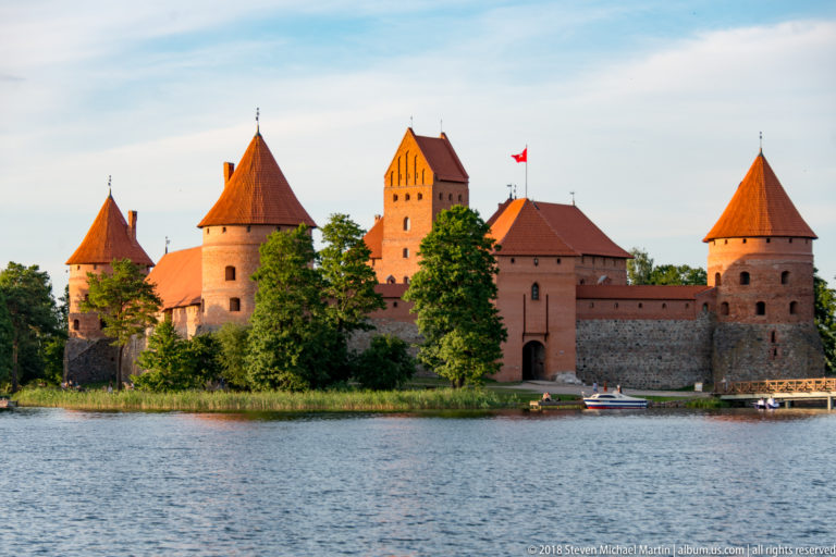 Trakai Island Castle across Lake Galvė, with red-brick towers, curtain walls, trees, and a small boat near shore.